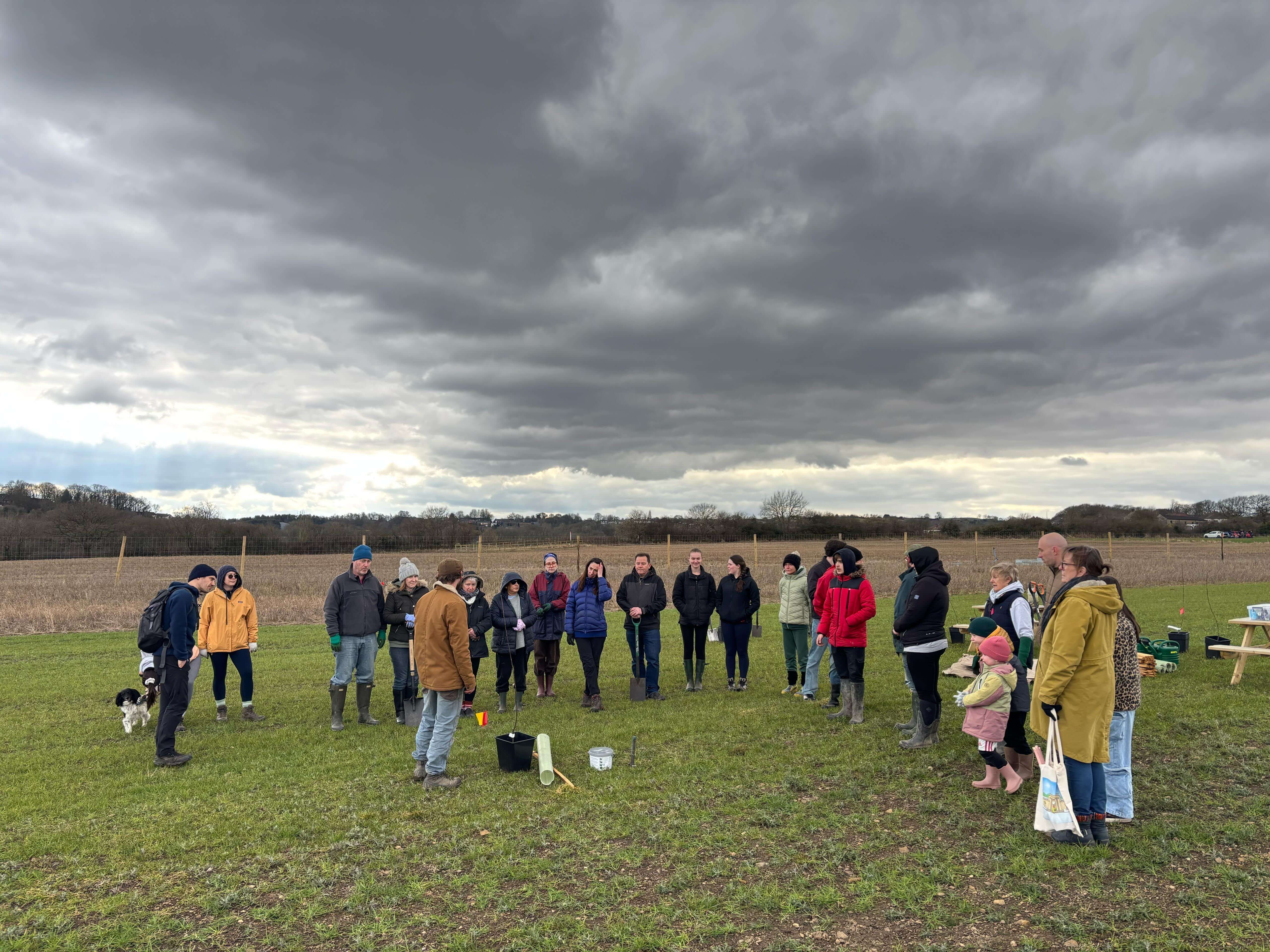 Members of the community gathering in a field on an overcast day to listen to our Patch Manager, Will, share how to plant a fruit tree in the ground